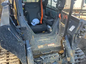 Close-up of a muddy John Deere skid steer loader with a white safety helmet inside the cab on a construction site.