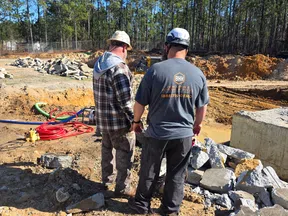 Two construction workers wearing helmets inspect rocky excavation site with trees and equipment nearby under clear blue sky.