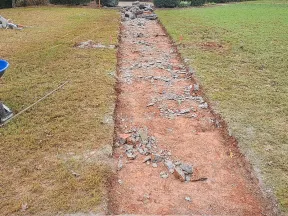 Excavated dirt path with scattered rocks and lawn tools, including a blue wheelbarrow and rake, on grass.