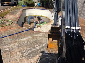 Workers repair and clean an empty swimming pool using hoses and heavy machinery on a sunny day.