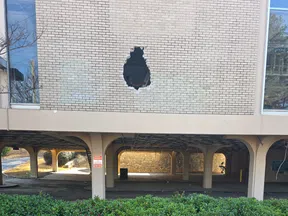 Brick wall with a large hole overlooking a parking area with columns and a green bush in front.