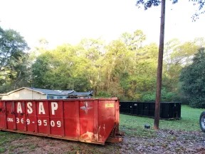 Large red dumpster with ASAP and phone number on grass near trees and a small house in the background.