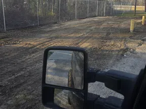 Side mirror view of a vehicle beside a dirt road with chain-link fence and tall leafless trees in the background.