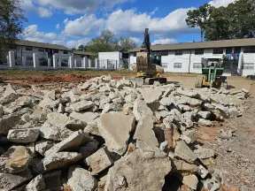 Construction site with broken concrete rubble in foreground and excavator and loader against residential buildings.