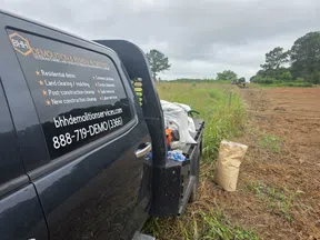 Black demolition service truck parked by a partly cleared grassy field with a driver in the cab on a cloudy day.