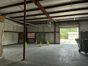 Empty industrial warehouse with concrete floor, exposed beams, green safety cones, and open loading dock door.