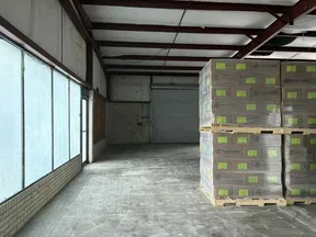 Interior of an empty warehouse with stacked pallets of boxes near a closed roll-up door and frosted windows.