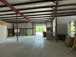 Empty industrial warehouse interior with concrete floor, steel beams, pallets, and open garage door letting in daylight.