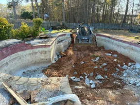 Partially demolished backyard pool being filled with dirt and rubble by a small bulldozer under clear daylight.