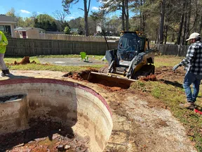 Construction workers use heavy machinery to excavate soil around an empty backyard pool during renovation.