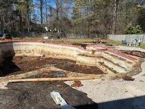Empty, dirty swimming pool with deteriorated walls and debris inside in a backyard surrounded by trees and a fence.
