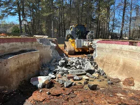 Bulldozer demolishing old concrete swimming pool with broken rubble inside on a sunny day.