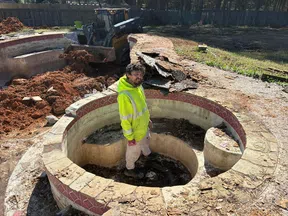 Worker in fluorescent jacket stands inside a circular stone fire pit under construction with construction equipment nearby