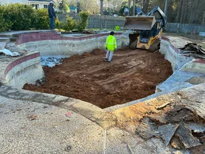 Construction workers excavate soil inside an empty cracked swimming pool using a small bulldozer.