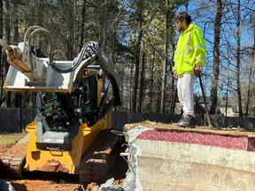 Construction worker in a neon jacket stands on a concrete slab near a John Deere excavator in a wooded area.