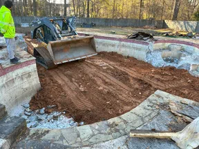 Construction of an empty swimming pool with compacted dirt and a skid steer loader inside, worker observes the site.
