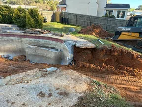 Partial demolition of an outdoor pool with exposed concrete, dirt piles, and a Deere excavator in a backyard.