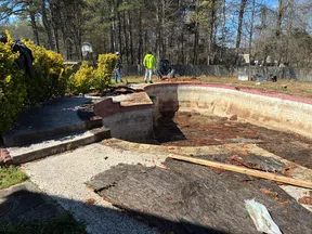 Empty, deteriorated in-ground swimming pool being renovated outdoors with workers in background and surrounding trees.