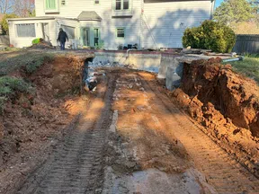 Deep excavation trench with exposed soil and tire tracks near a white residential house under construction.