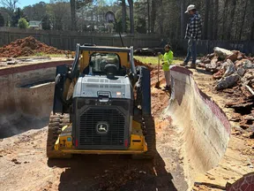 John Deere skid steer working in an empty old pool with two workers managing demolition and dirt removal outdoors.