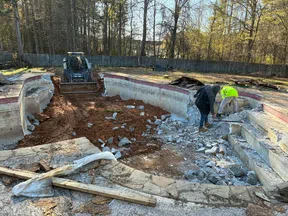 Construction workers and excavator demolishing an old concrete swimming pool surrounded by trees and a wooden fence