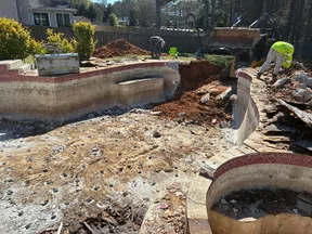 Two workers demolishing an old backyard swimming pool during daytime with heavy machinery and debris around.