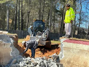 Construction workers demolishing concrete foundation with bulldozer and tools in outdoor site with trees.