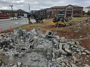 Construction site with rubble, exposed rebar, and heavy machinery near urban apartment buildings under cloudy sky.