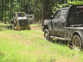 Black pickup truck towing a bulldozer through a grassy forest clearing on a sunny day