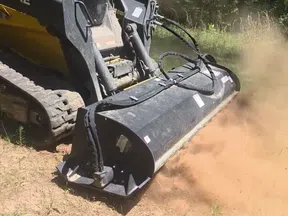 Tracked skid steer with brush cutter attachment clearing dry grass and dirt, creating dust cloud in a sunny outdoor setting.