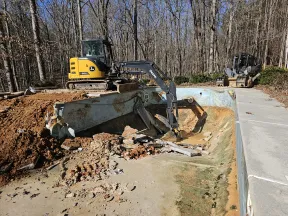 Mini excavator demolishing an old concrete pool surrounded by leafless trees on a sunny day.