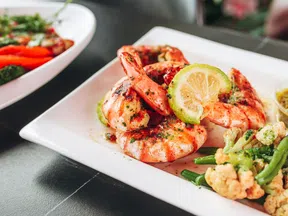 Grilled shrimp garnished with lemon slice and herbs served with steamed vegetables on a white plate.