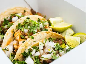 Three street tacos with meat, chopped onions, cilantro, and lime wedges in a white takeout box