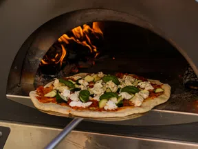 Fresh pizza topped with basil, cheese, and vegetables being placed into a wood-fired outdoor oven