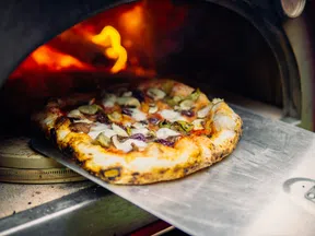 Freshly baked pizza with vegetables and cheese being removed from a wood-fired oven with visible flame.