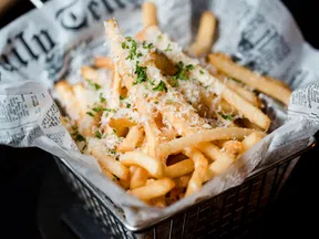 Basket of golden French fries topped with grated cheese and fresh green herbs on newspaper-style paper.
