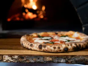 Wood-fired pizza with melted cheese and basil on a wooden peel in front of a glowing brick oven fire.