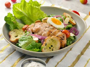 Grilled chicken salad with lettuce, cherry tomatoes, boiled egg, croutons, and creamy dressing on a striped cloth.