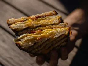 Close-up of a grilled cheese sandwich with melted cheese and bacon held over a wooden table.
