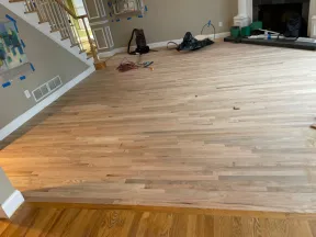 Freshly installed light oak hardwood flooring in living room under renovation with staircase and fireplace.