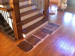 Wood floor repair patches in front of wooden staircase with railing in a residential interior.