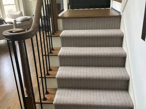 Carpeted stairs with patterned beige runner and wooden handrail leading to a bright living room area.