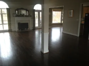 Spacious empty living room with dark hardwood floors, fireplace, large windows, and white pillar in center.