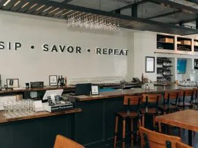 Modern bar interior with wooden stools, wine glasses, and wall text reading SIP SAVOR REPEAT above counter.