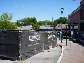 Construction site fenced with black Dakota-branded barriers in a sunny outdoor shopping area with trees and buildings.
