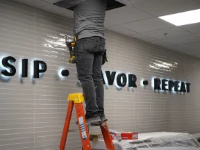 Worker on a ladder installing illuminated wall letters reading SIP VOR REPEAT on tiled wall.