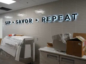 Illuminated SIP SAVOR REPEAT sign on tiled wall in room with covered furniture and unpacked boxes.