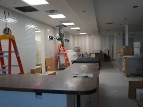 Under-construction interior with ladders, tools, boxes, and a worker installing fixtures in a commercial space.