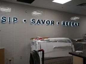 Illuminated wall sign reading SIP • SAVOR • REPEAT over a kitchen countertop with covered appliances and prep area.