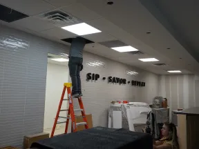 Worker on ladder installing ceiling tiles in a room with tiled walls displaying 'SIP SAVOR RELAX' sign.